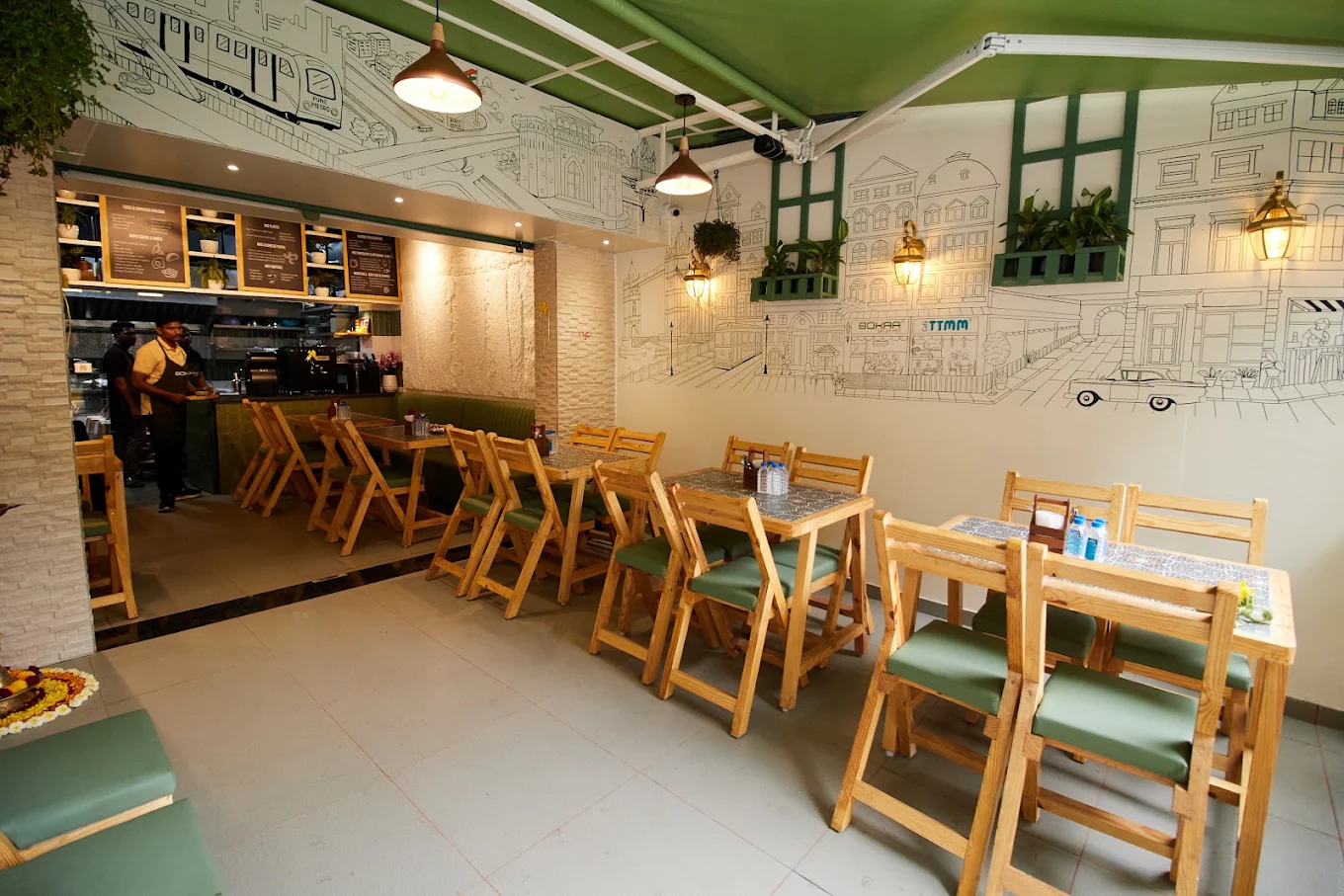 Inside Bokaa: pine folding chairs and green banquette seating under a black line drawing of the Pradhikaran streetscape